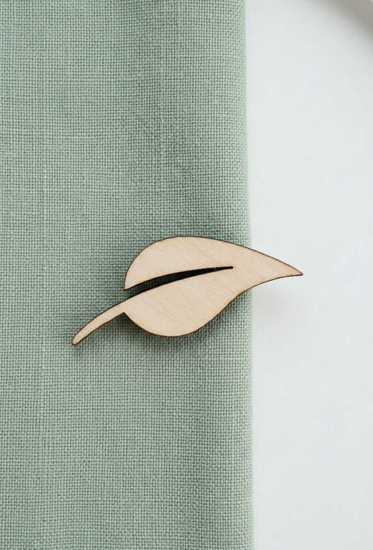 A light natural wood leaf-shaped lapel pin with a dark center slit, pinned to a sage green textured fabric against a white background.