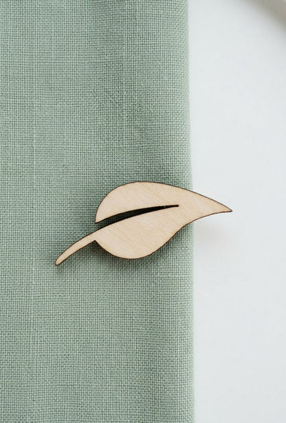 A light natural wood leaf-shaped lapel pin with a dark center slit, pinned to a sage green textured fabric against a white background.