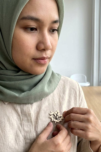 A close-up lifestyle shot of a woman wearing a sage green hijab and a light beige linen top, adjusting a small, laser-cut wooden leaf-shaped brooch on her chest.