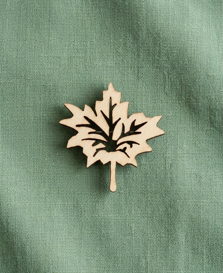 A laser-cut wooden maple leaf brooch with a natural light wood finish, displayed on a textured sage green fabric background.