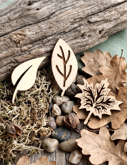 Three unfinished light wood leaf-shaped brooches with engraved leaf vein details, arranged on a rustic surface with dried oak leaves, moss, small stones, and a piece of driftwood.