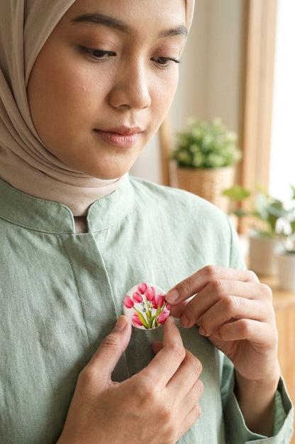 A woman wearing a light beige hijab and a sage green linen top adjusts a small pink and white flower pin on her chest. The background shows a softly blurred indoor setting with warm lighting and potted plants.