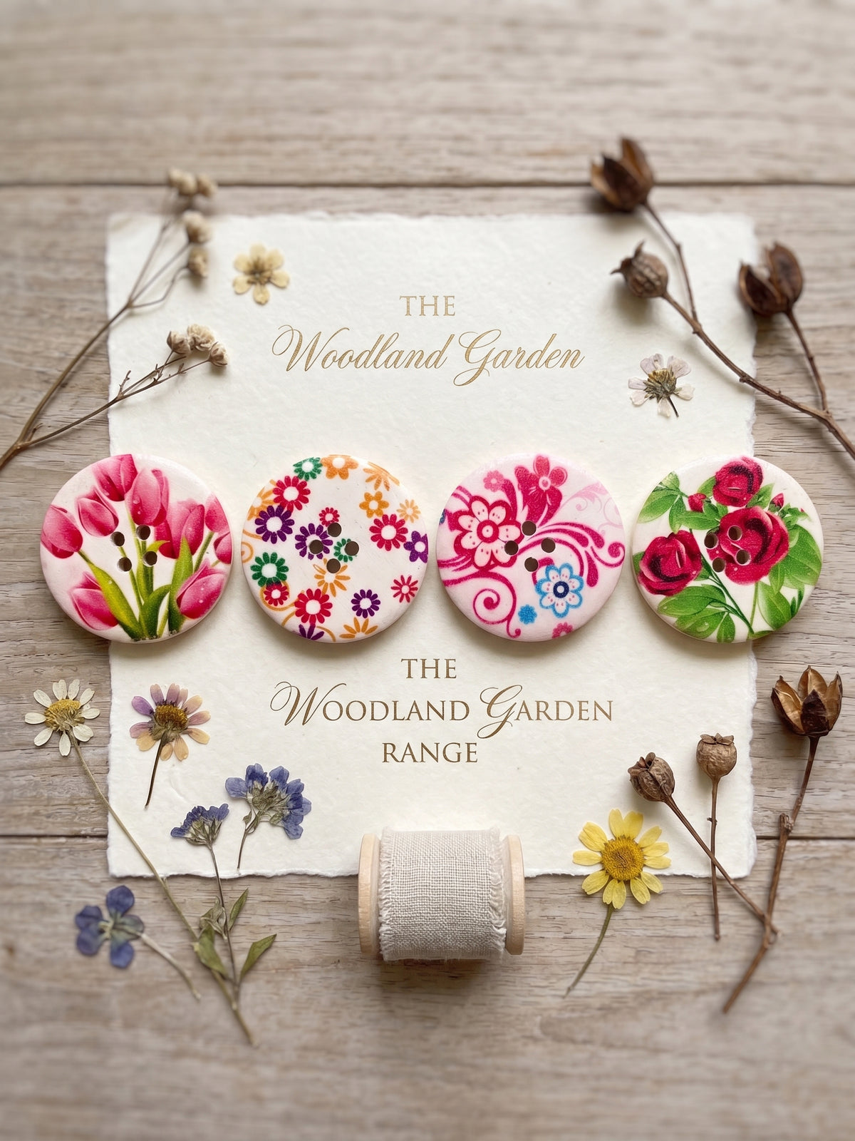 Four round floral buttons displayed on a cream paper card with dried flowers and a spool of thread around it. The card reads 'The Woodland Garden Range.