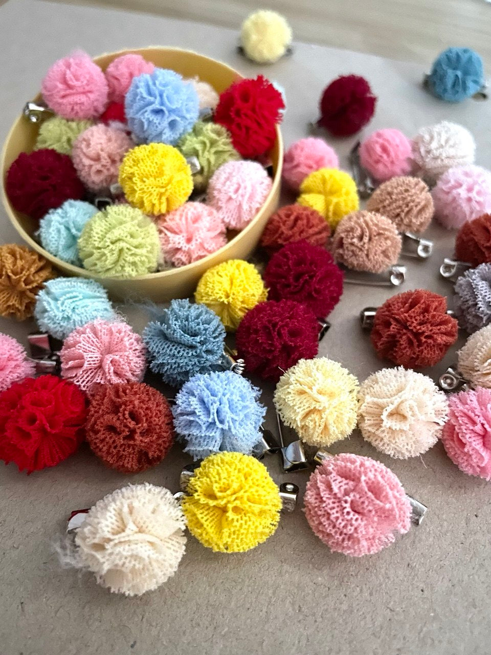Colorful pom-pom mini brooches on a gray surface with a bowl of additional pom-poms.