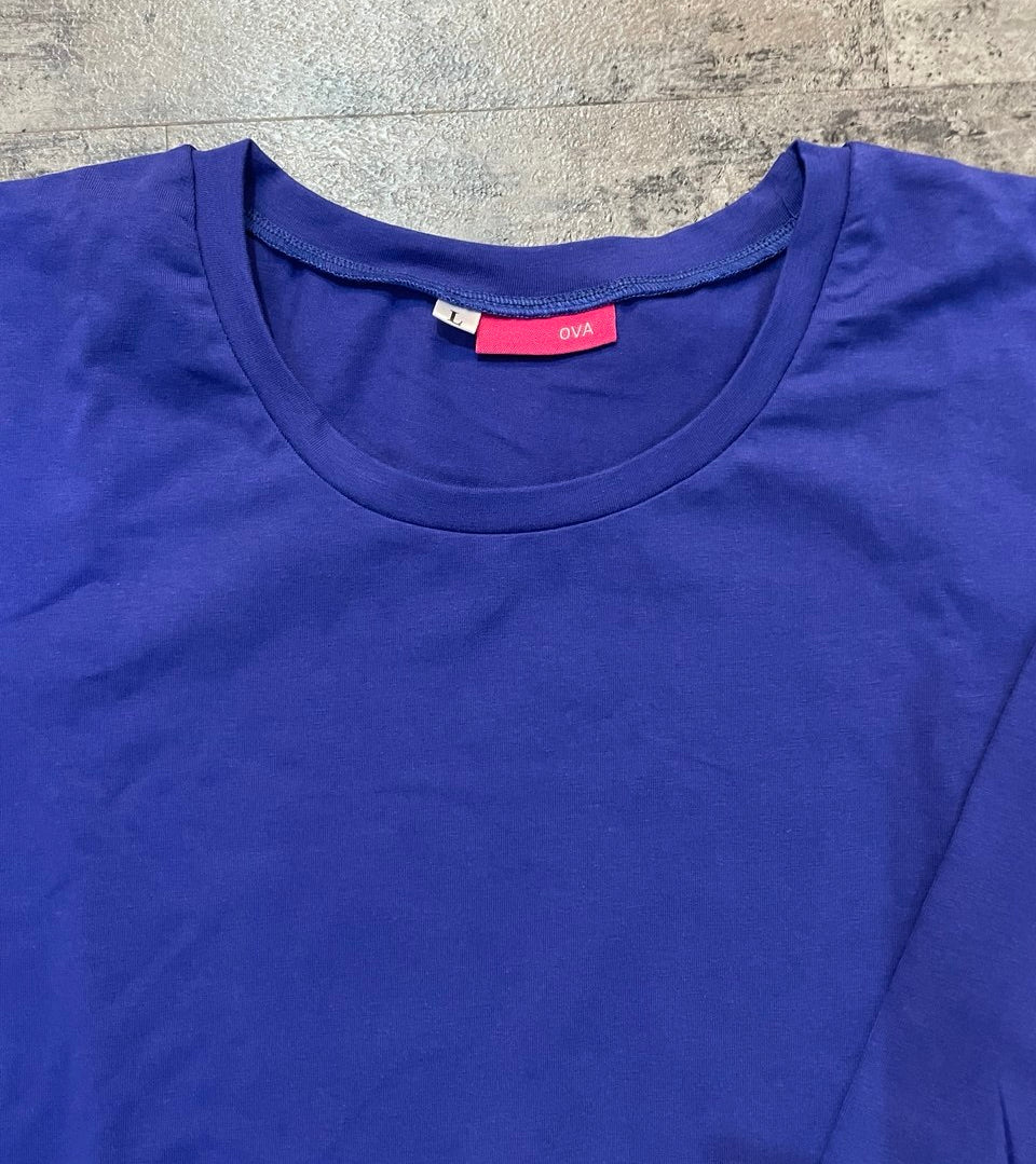 A close-up of a royal blue crewneck t-shirt laid flat on a gray textured surface, showing a pink inner neck label with the brand name 'ova'.