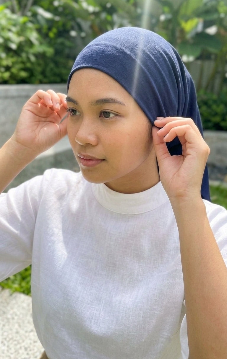 A woman wearing a navy blue hijab and a white mock-neck top, adjusting the edges of her hijab with both hands in an outdoor setting with green foliage in the background.