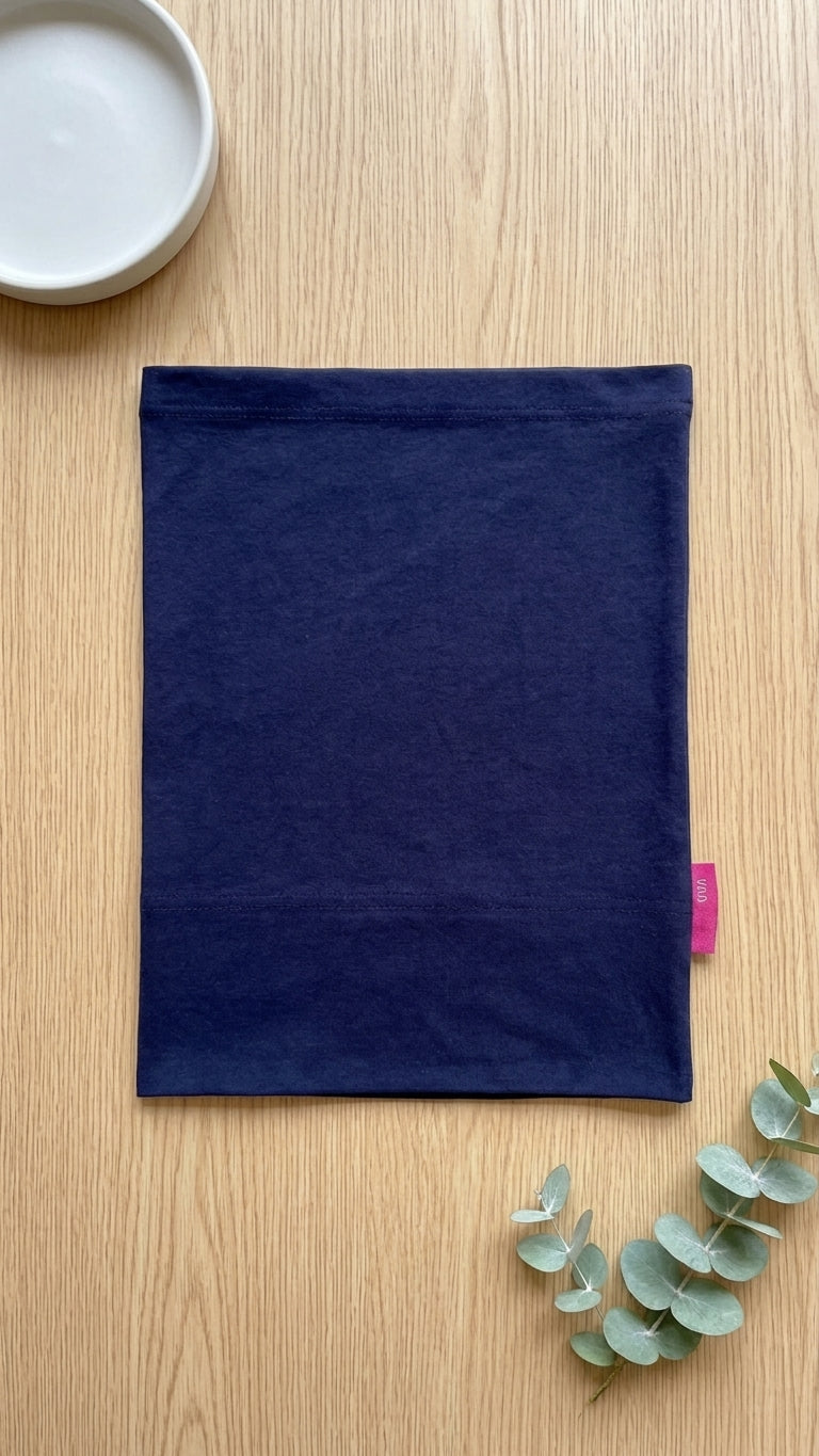 A navy blue fabric sleeve or pouch laid flat on a light wood tabletop, with a small pink tag visible on the right edge. A white dish is partially visible in the top-left corner and eucalyptus leaves are placed in the bottom-right corner.