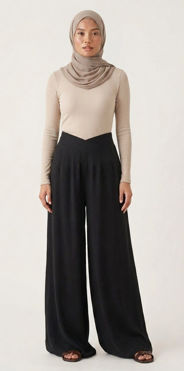 A woman modeling a beige long-sleeve top and black wide-leg pants with a taupe hijab, standing against a plain off-white studio background.