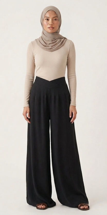A woman modeling a beige long-sleeve top and black wide-leg pants with a taupe hijab, standing against a plain off-white studio background.