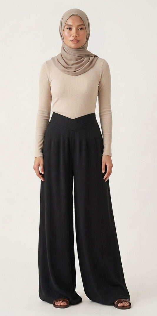 A woman modeling a beige long-sleeve top and black wide-leg pants with a taupe hijab, standing against a plain off-white studio background.