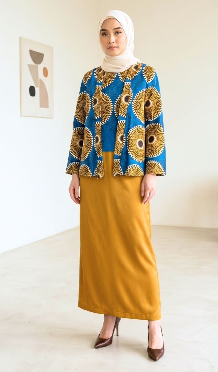 A woman wearing a two-piece outfit consisting of a blue and gold patterned long-sleeve top and a mustard yellow maxi skirt, paired with a white hijab and brown heels, standing in a minimalist room.
