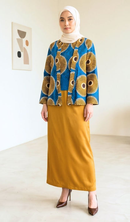 A woman wearing a two-piece outfit consisting of a blue and gold patterned long-sleeve top and a mustard yellow maxi skirt, paired with a white hijab and brown heels, standing in a minimalist room.