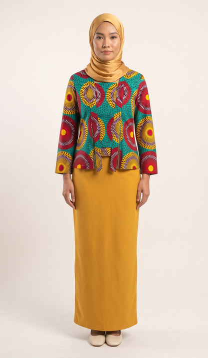 A woman modeling a two-piece modest outfit consisting of a mustard yellow skirt and a matching hijab, paired with a teal long-sleeve top featuring a vibrant red, yellow, and orange circular pattern.