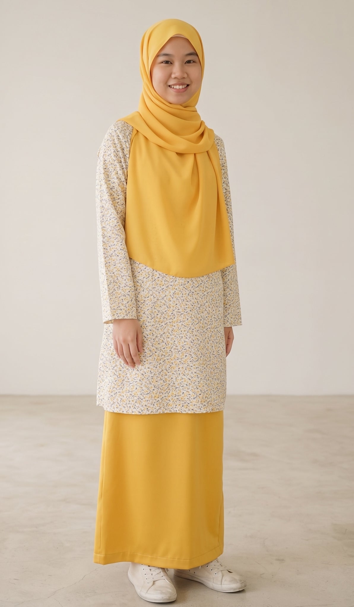 A woman wearing a modest outfit consisting of a mustard yellow hijab, a long-sleeved tunic with a cream and yellow floral pattern, and a matching solid mustard yellow skirt, standing against a neutral beige background.