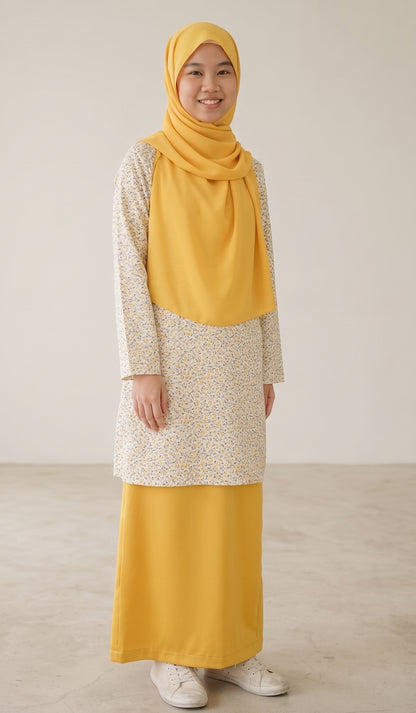 A woman wearing a modest outfit consisting of a mustard yellow hijab, a long-sleeved tunic with a cream and yellow floral pattern, and a matching solid mustard yellow skirt, standing against a neutral beige background.