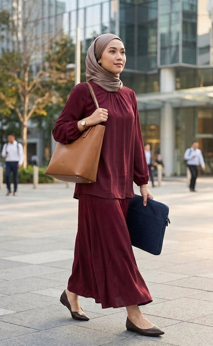 A woman wearing a matching burgundy long-sleeve maxi dress and beige hijab walks outdoors in a modern city setting, carrying a large brown tote bag and a black laptop.