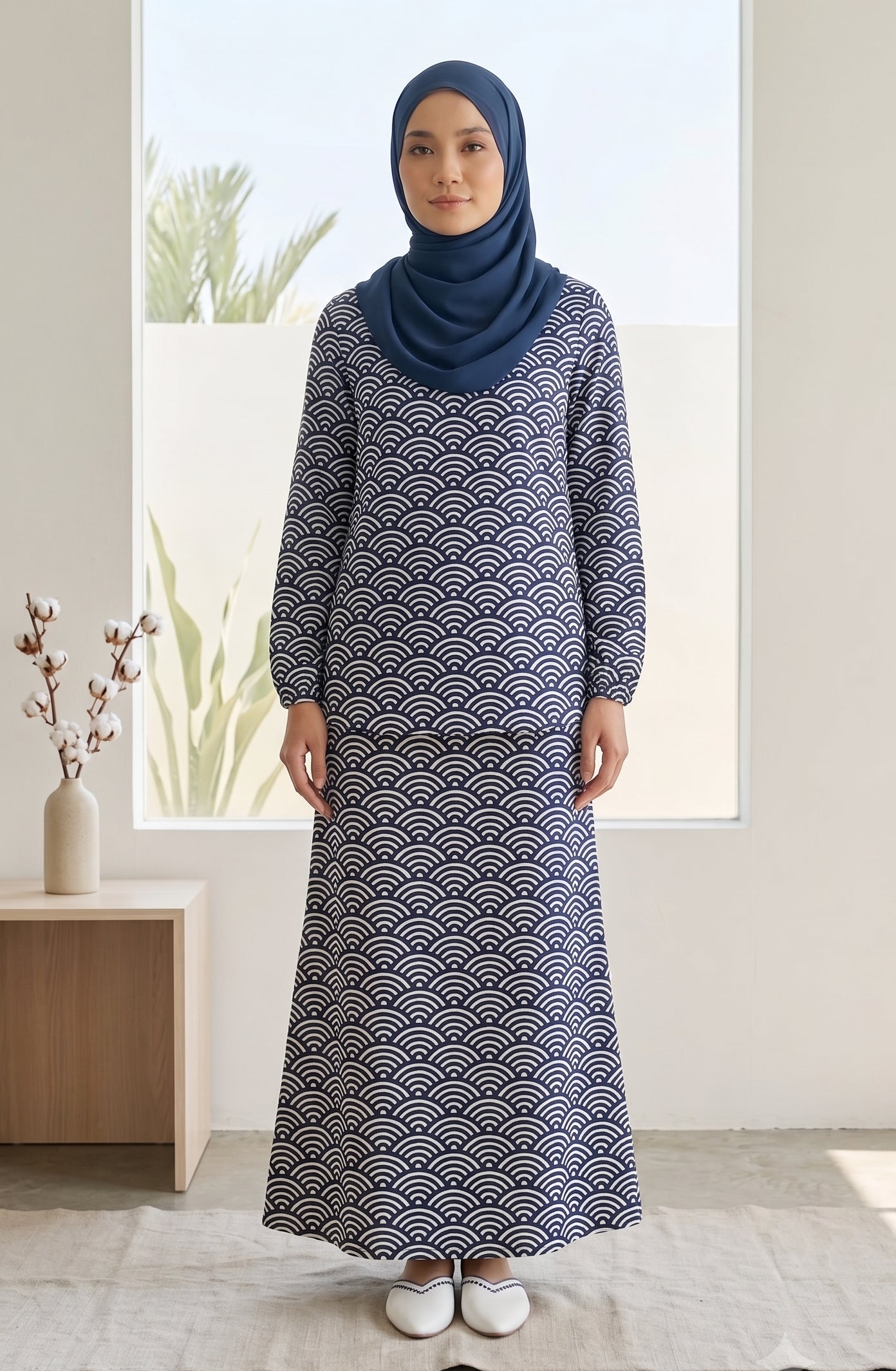 A woman wearing a long-sleeved navy blue maxi dress with a white repeating wave pattern and a matching navy blue hijab, standing in a bright, modern room.