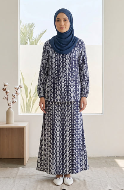 A woman wearing a long-sleeved navy blue maxi dress with a white repeating wave pattern and a matching navy blue hijab, standing in a bright, modern room.