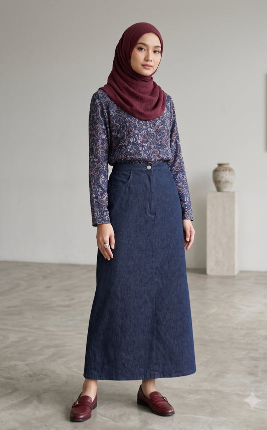 A woman wearing a dark blue denim maxi skirt, a patterned long-sleeve top, and a burgundy hijab, standing in a minimalist studio setting.