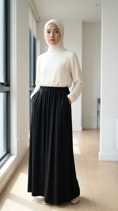A woman wearing a cream mock-neck long-sleeve top and a long black skirt with side pockets, paired with a light beige hijab, standing in a bright, modern hallway with wooden floors.