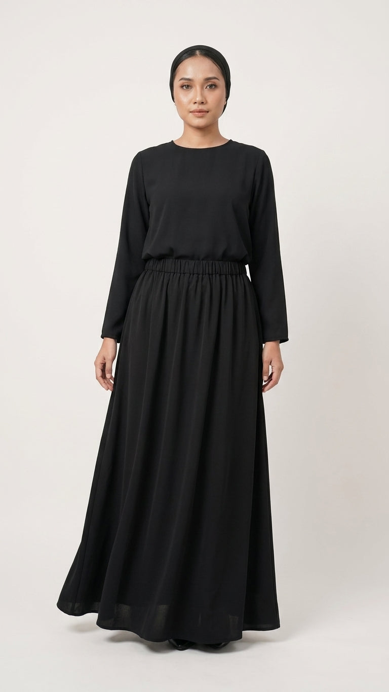 A woman modeling a long-sleeved black maxi skirt and blouse with a high round neckline and an elastic waist, standing against a light gray studio background.