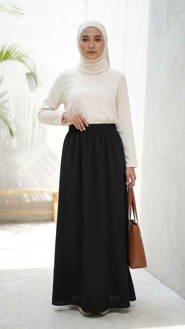 A woman wearing a white mock-neck long-sleeve top and a long black skirt, paired with a white hijab and carrying a brown leather tote bag, standing in a bright, modern indoor setting with a white wall.