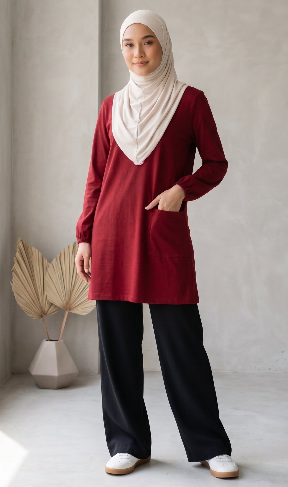 A woman wearing a long-sleeve burgundy tunic with a round-neck and a patch pocket at right, paired with wide-leg navy pants and a cream hijab, standing in a minimalist studio setting.