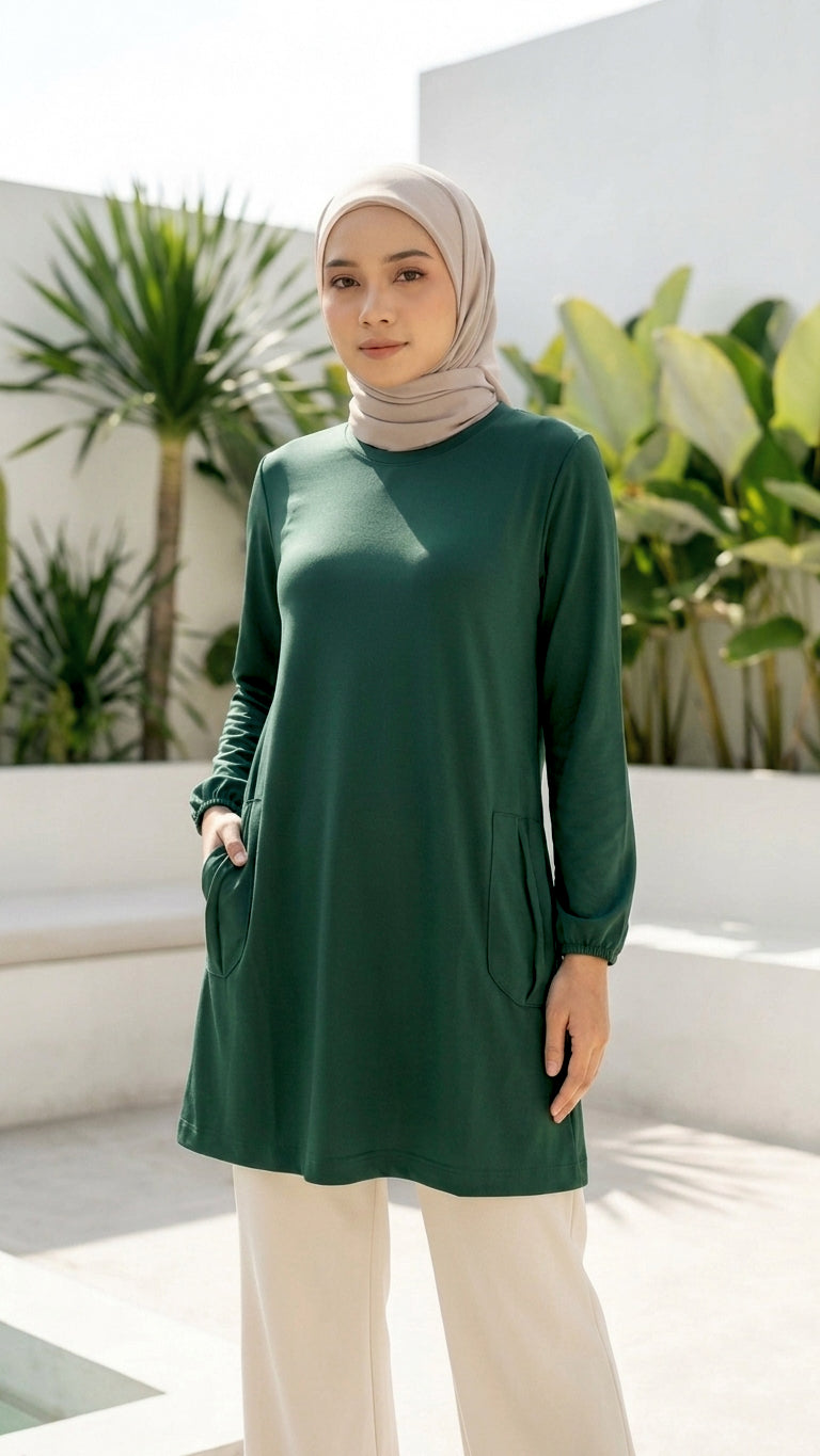 A woman wearing a dark green long-sleeve tunic with side pockets, a light beige hijab, and cream wide-leg trousers, standing in a modern outdoor patio setting with white walls and green plants.