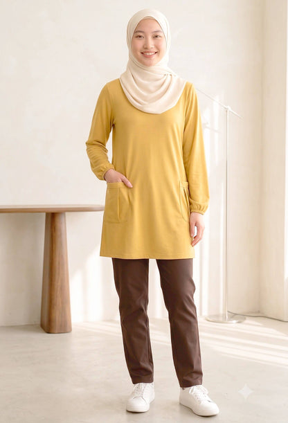 A woman wearing a mustard yellow long-sleeve tunic with side pockets, brown pants, and a cream hijab, standing in a bright, modern room.