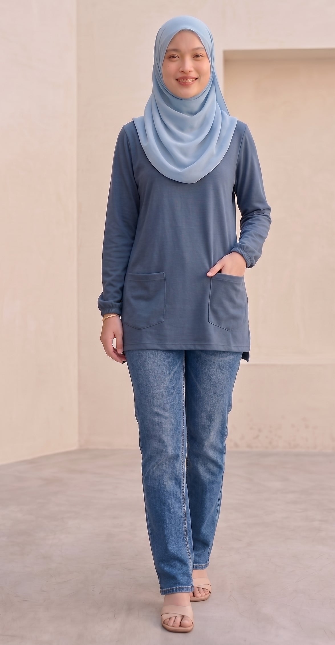 A woman wearing a dusty blue long-sleeve tunic with front pockets, blue jeans, and a light blue hijab, standing in a minimalist beige-walled indoor setting.