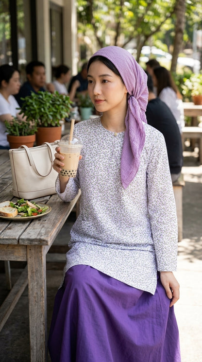 model wearing a long blouse in lilac and pruple skirt with a headscarf with a boba tea on her right hand and cafe background